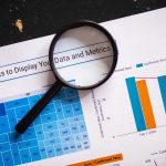 Lupa sobre gráficos e dados financeiros impressos, representando análise e revisão regulatória.