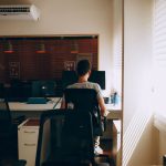 Homem trabalha diante de um computador em um escritório iluminado pela luz que entra pelas persianas.