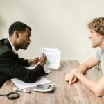 Consultor financeiro conversa com um cliente e analisa documentos em uma mesa de escritório.